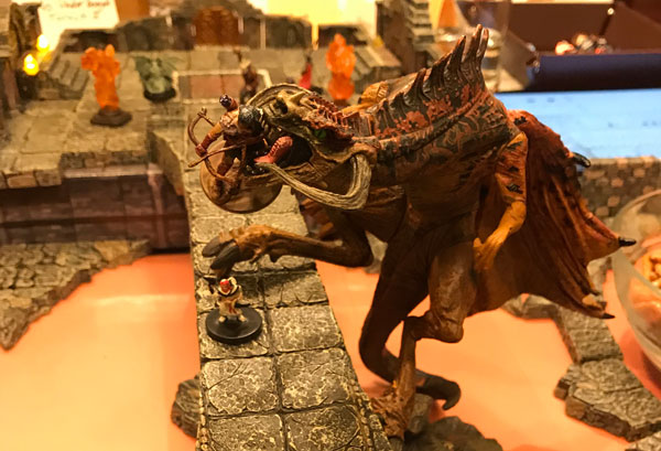 A dragon holds a miniature in its jaws as it climbs a stone bridge crossing a river of magma, all created with Dwarven Forge terrain.